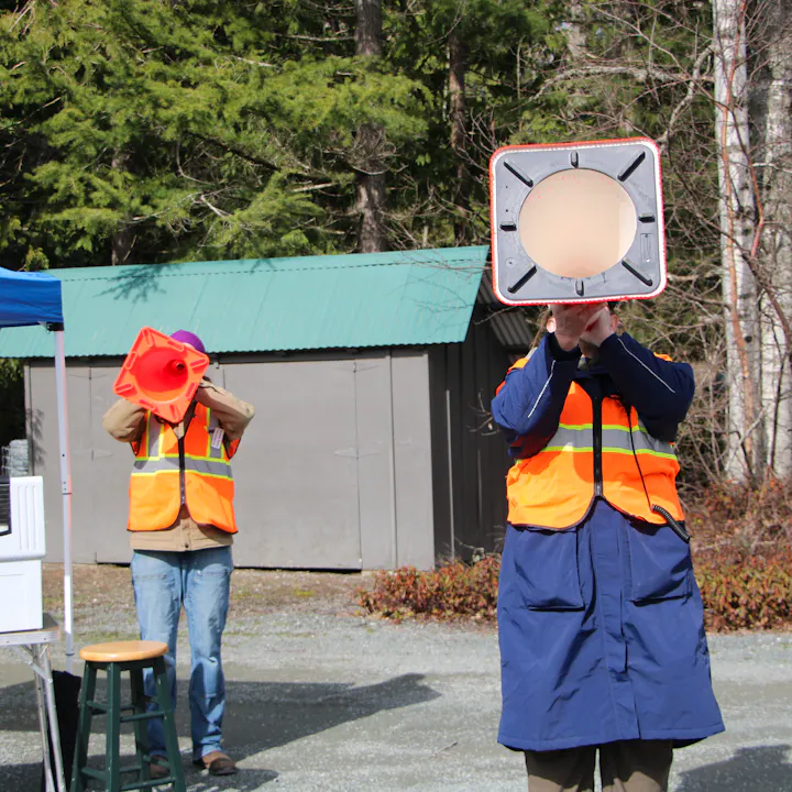 Two individuals in safety vests hold megaphones in a wooded area, with a green roof structure in the background.