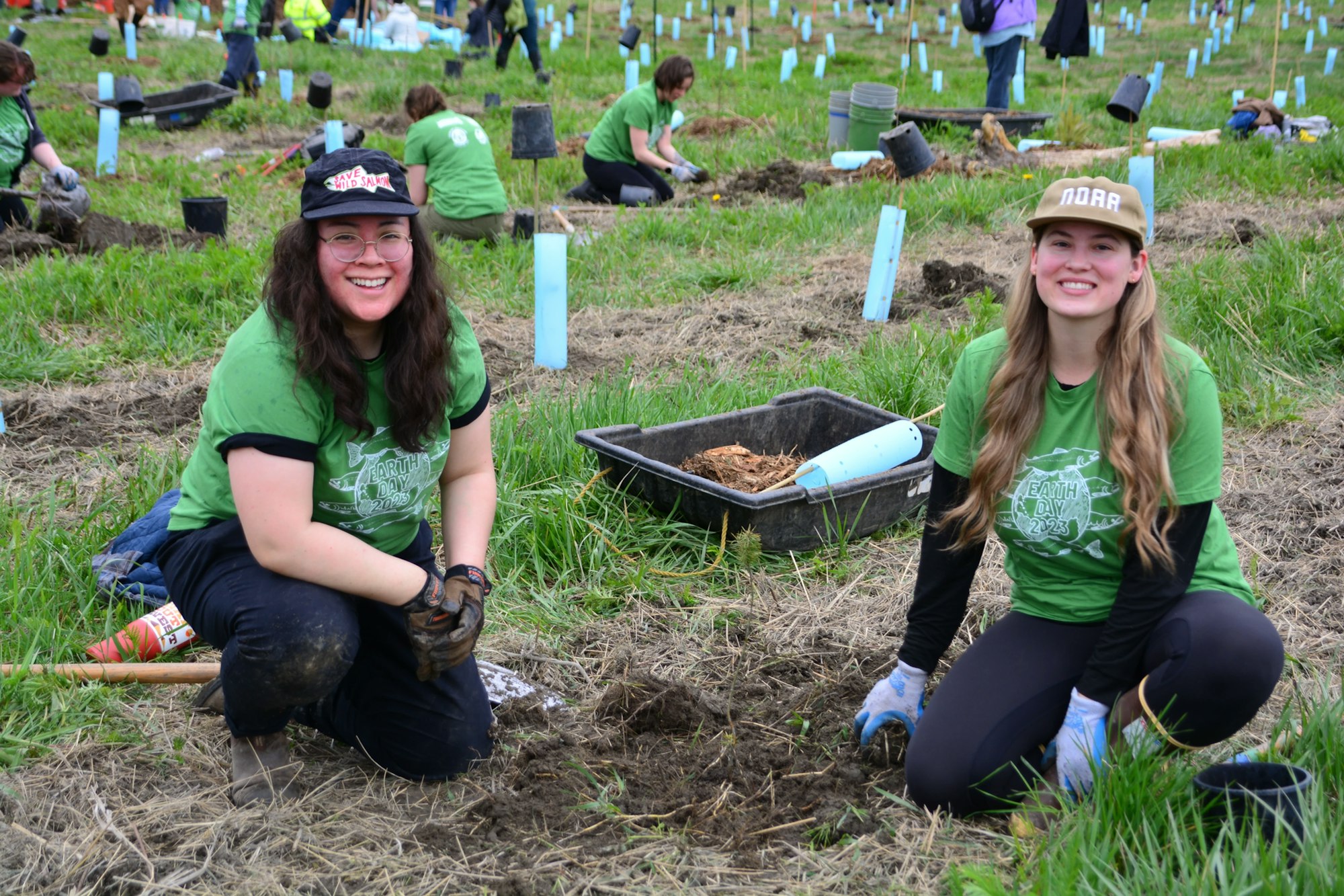 Two people planting in a field with others, wearing green "Earth Day 2023" shirts.