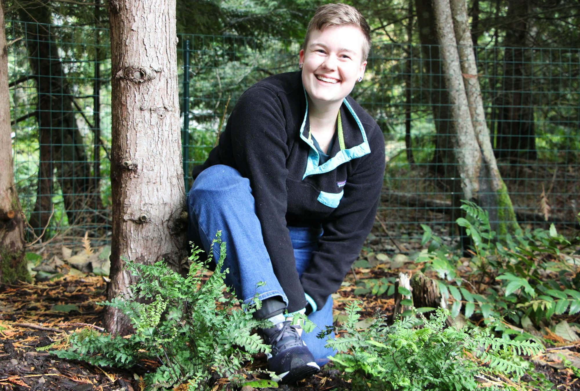 A person smiling, kneeling with a sneaker, in a forest setting with ferns and trees around.