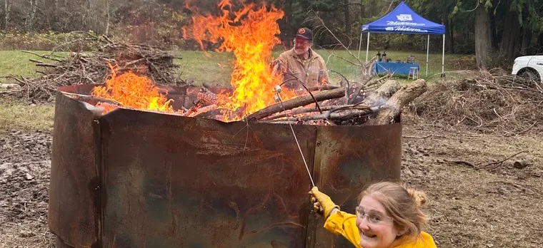 A person in a yellow jacket is smiling by a large fire pit with flames, while another person works nearby in a natural setting.