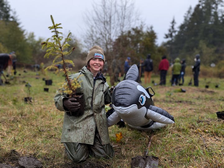 A person kneels in a muddy field with a small tree seedling and an inflatable orca, surrounded by others in the background.