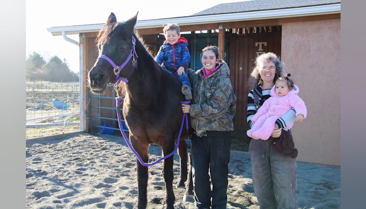 Two adults and two children with a horse in a sandy area near a building.