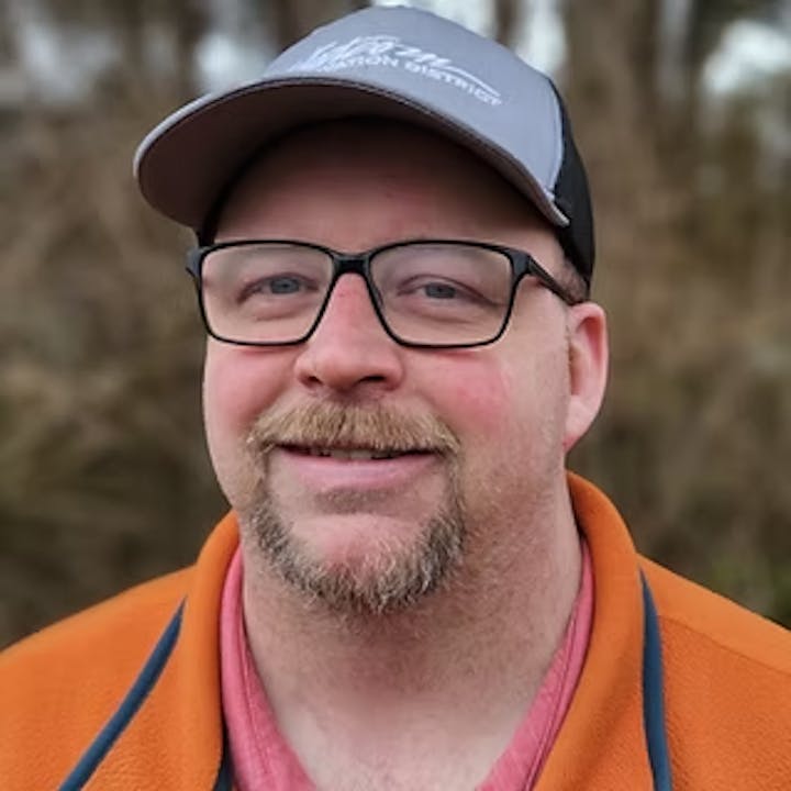 A person wearing glasses and a cap, smiling, with an outdoor background.