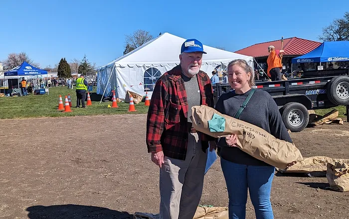 Two people standing outside near a tent, one holding a wrapped package, with a trailer in the background.