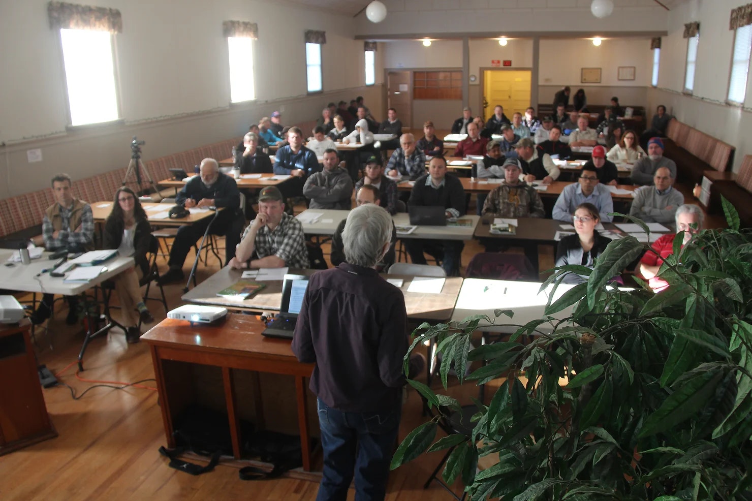 People in a classroom setting listening to a speaker, with desks, laptops, and a plant in the foreground.