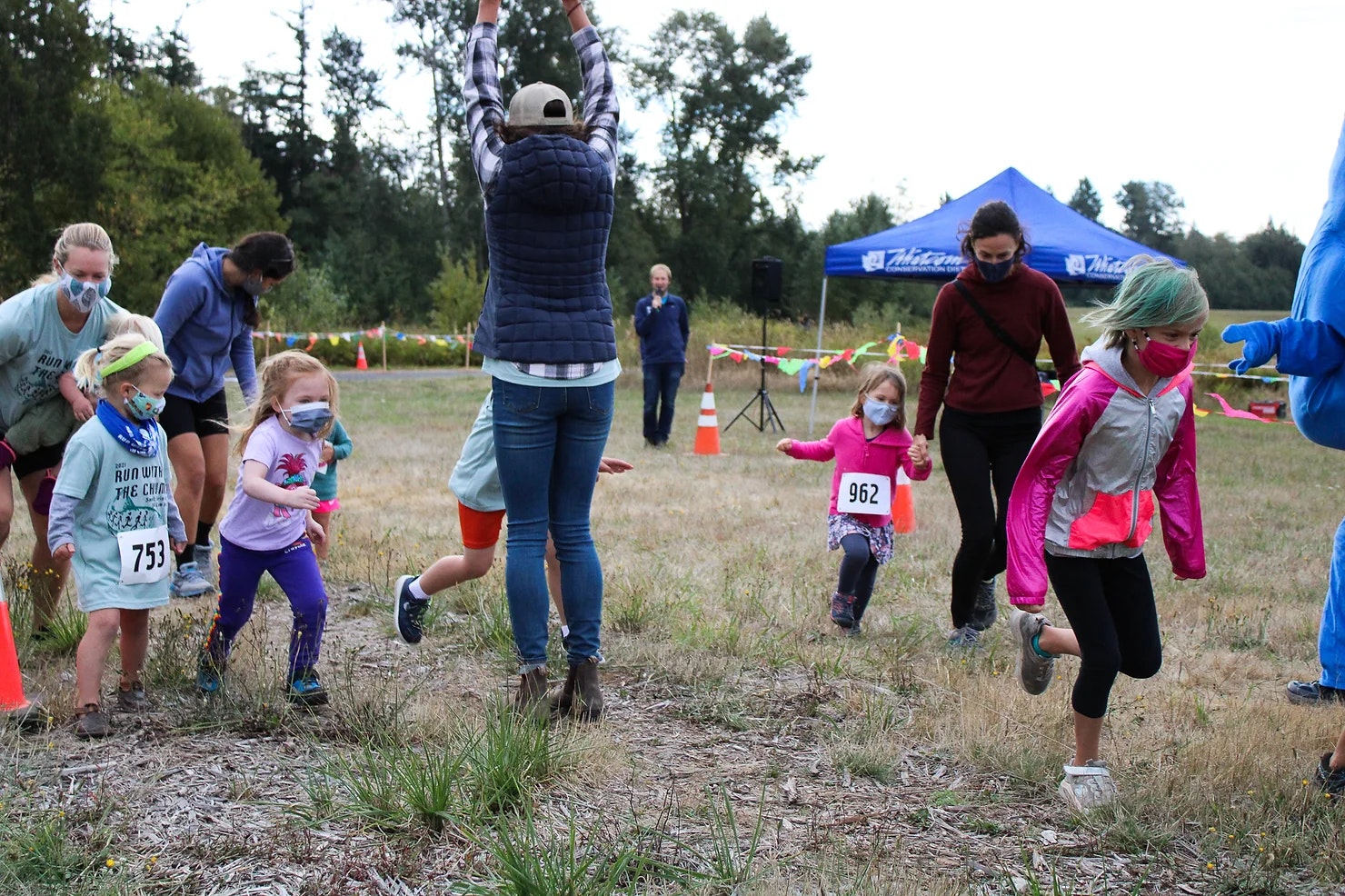 Children running in a race, wearing masks and numbered bibs, with adults supervising in a grassy outdoor area.