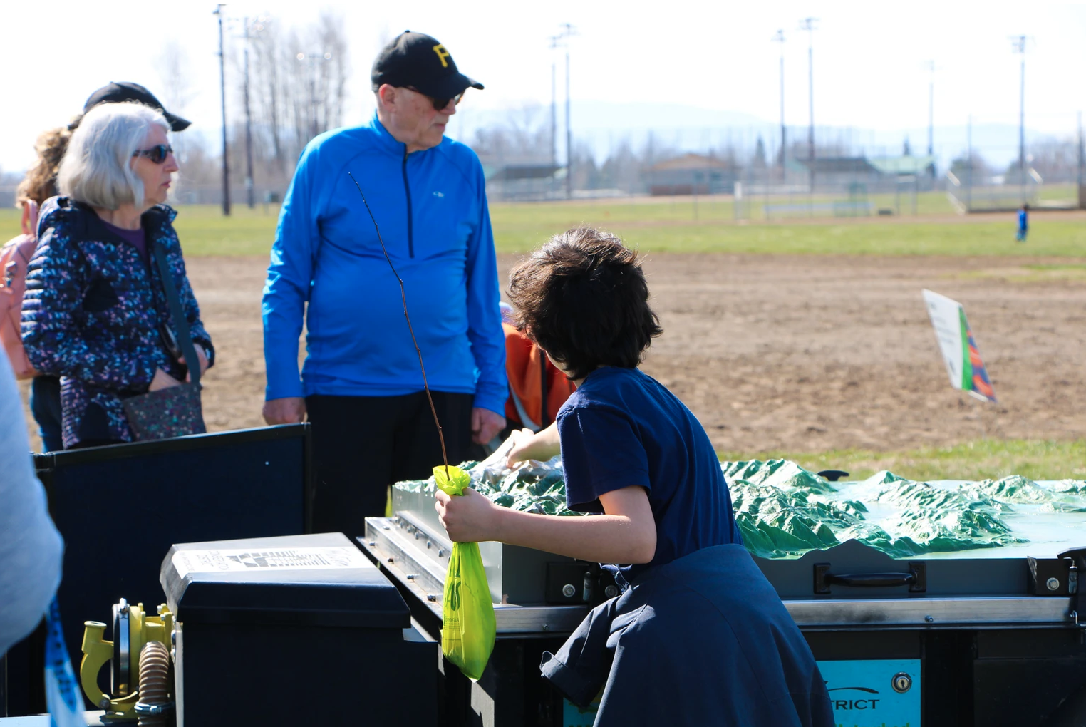 People gathered outdoors, observing a table with a green model. One person holds a yellow bag.