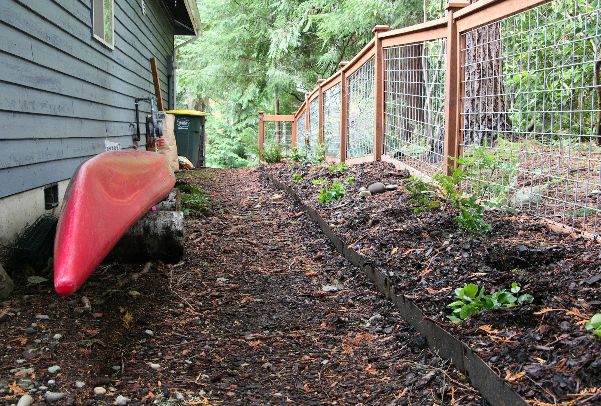 A red kayak is stored beside a house with a garden bed and wire fence bordering a wooded area.