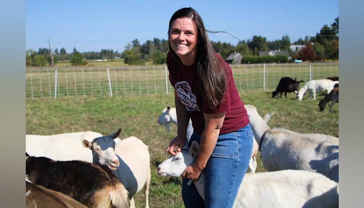 A person smiling in a field, surrounded by goats.