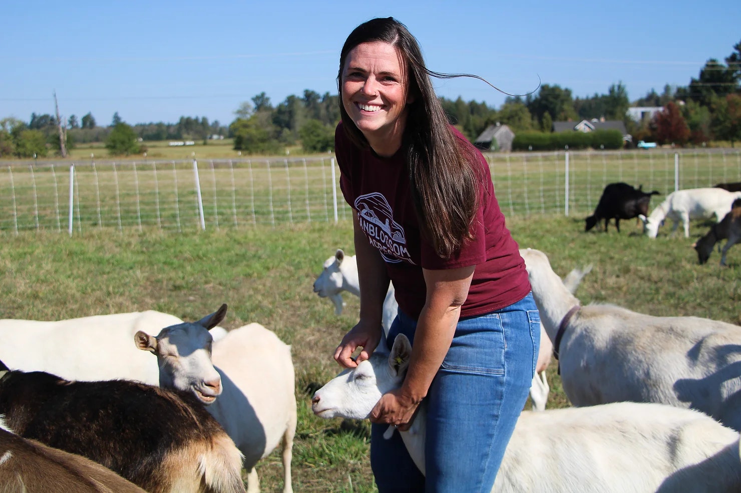 A person smiling in a field, surrounded by goats.