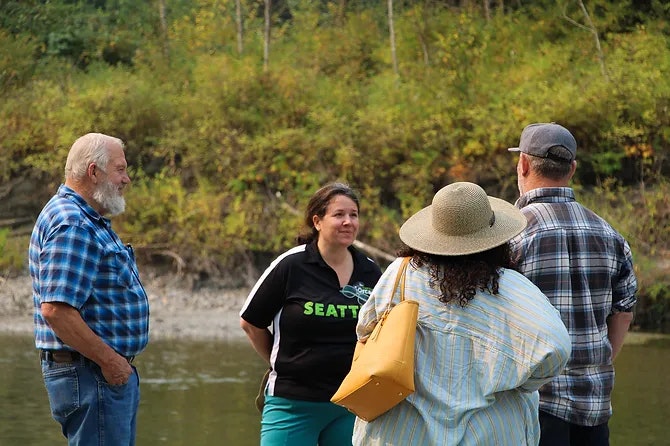 Four people chatting near a wooded riverside; one wears a "Seattle" shirt.