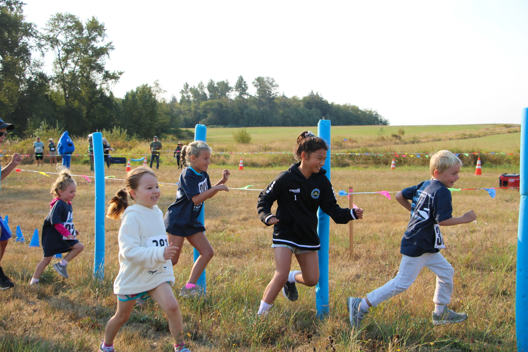 Children running in a field race, smiling and energetic, with colorful markers and a sunny outdoor setting.