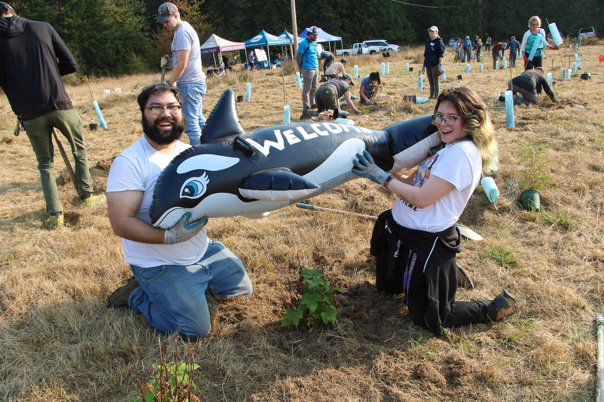 Two people holding an inflatable orca with "WELCOME" on it, at a tree planting event outdoors.