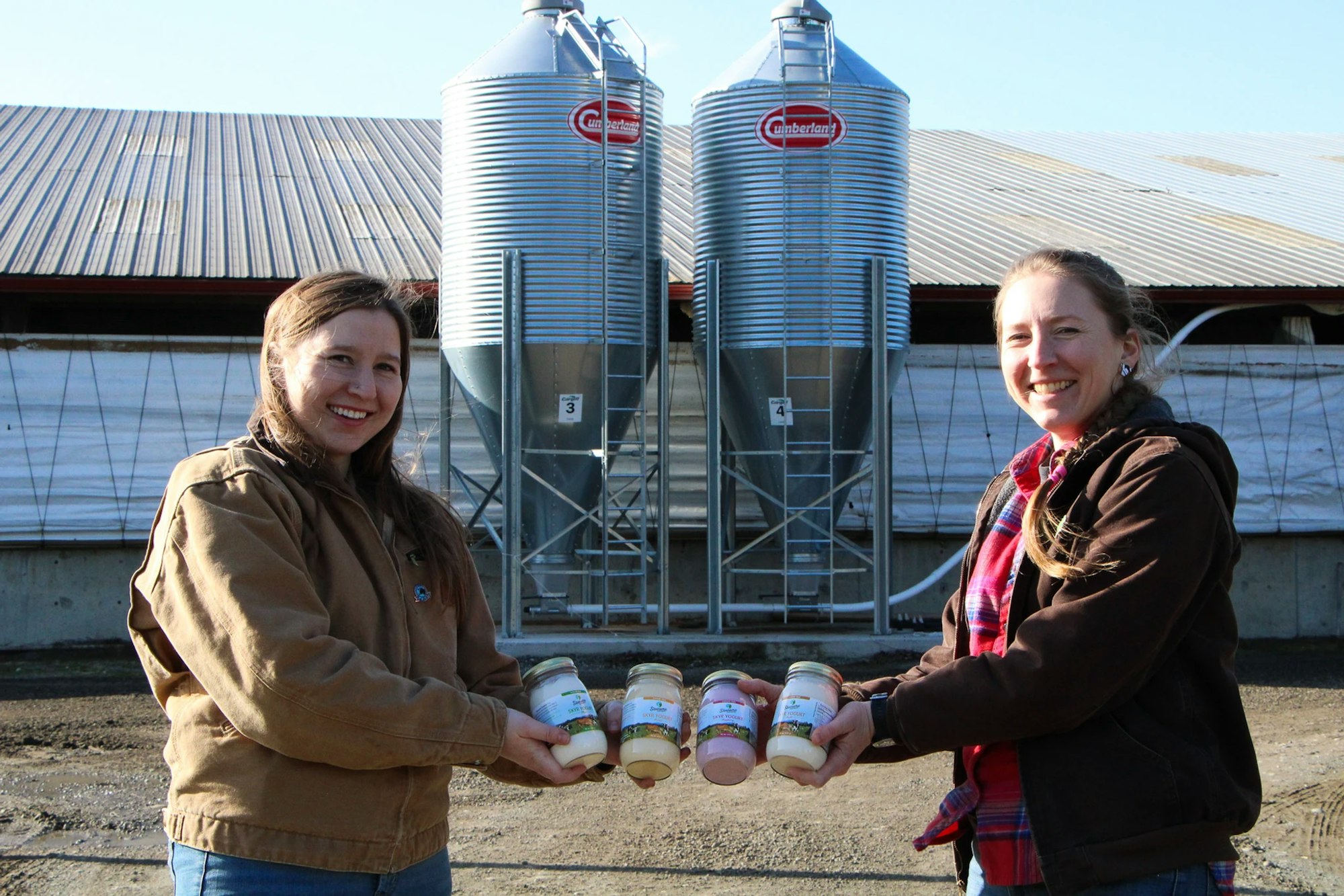 Two people smiling and holding jars, standing in front of large metal silos.