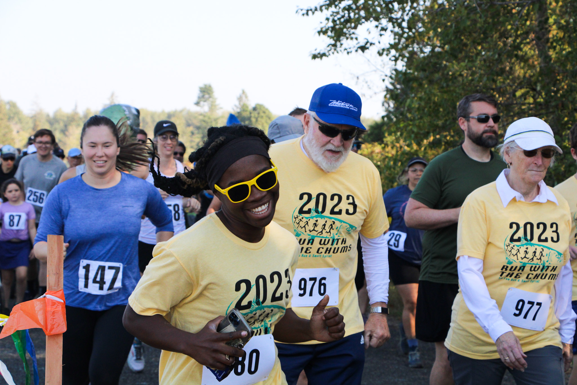 A crowd of happy participants in a 2023 run event, wearing T-shirts and race numbers, moving down a tree-lined path.