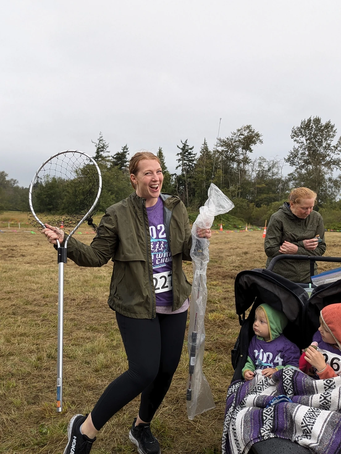 A person is happily holding a net and plastic-wrapped item, standing near two children in a stroller outdoors.
