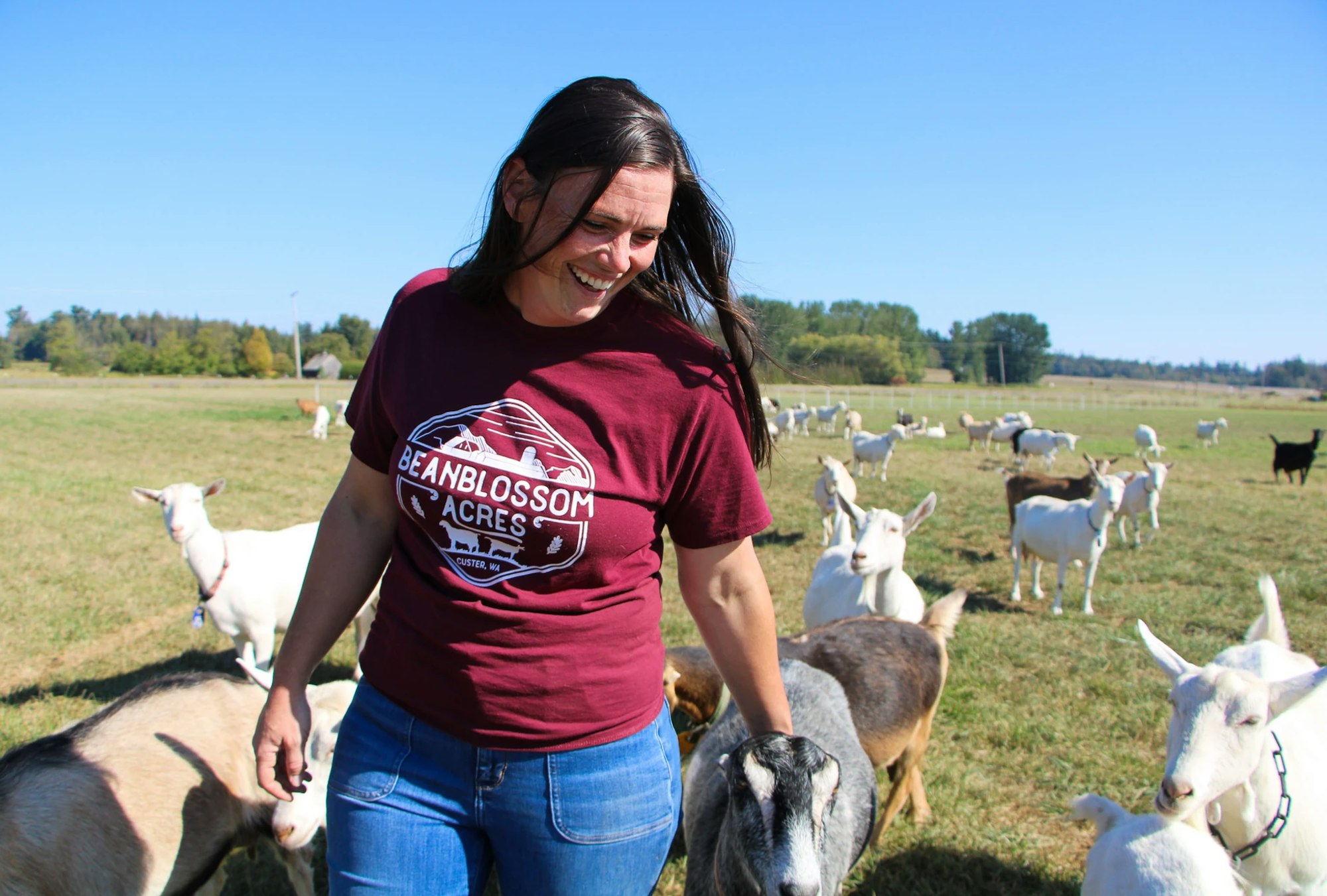A person smiling in a field with many goats, wearing a "Beanblossom Acres" shirt.