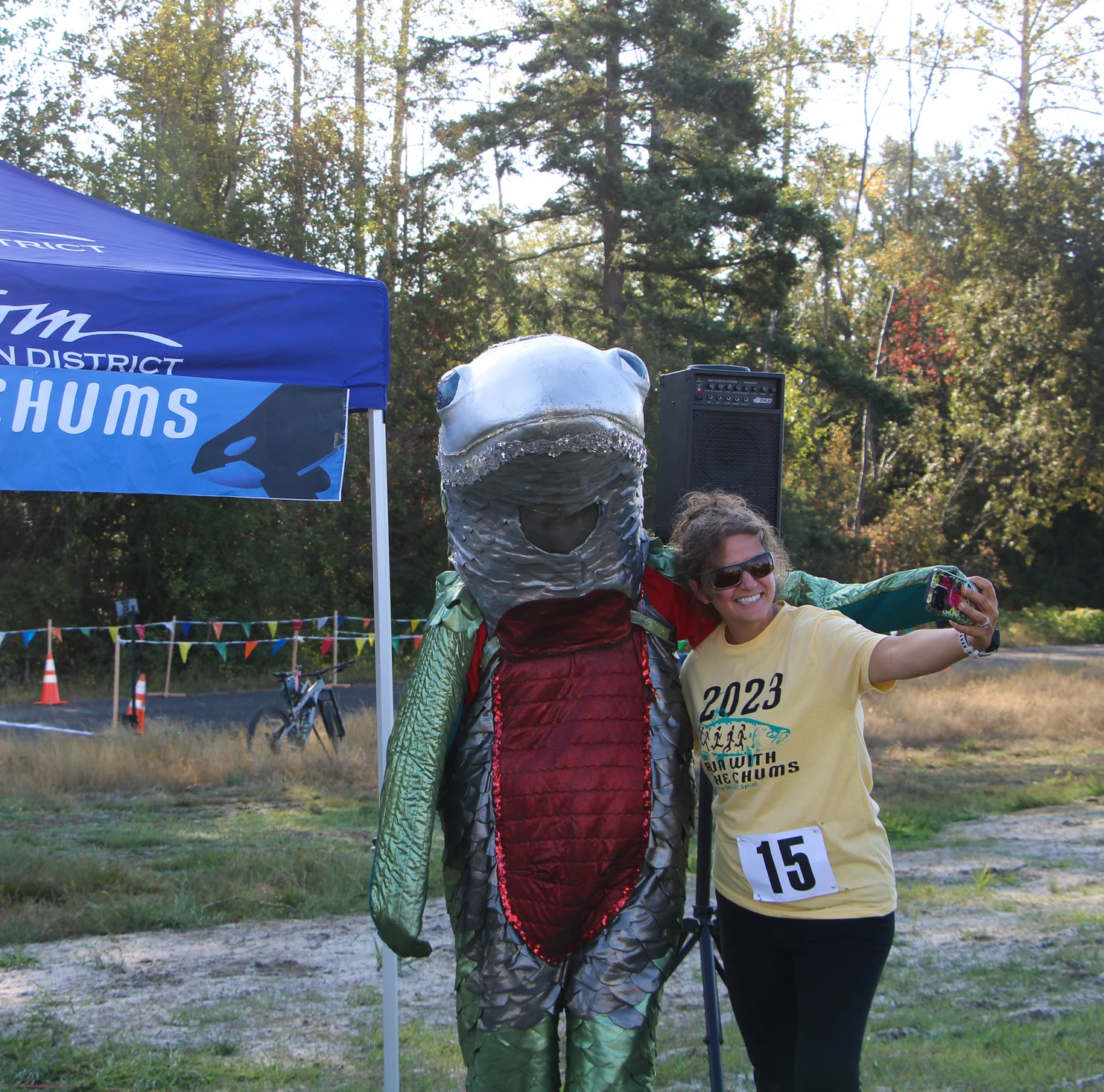 Person in a fish costume and a woman take a selfie under a tent at an outdoor event.