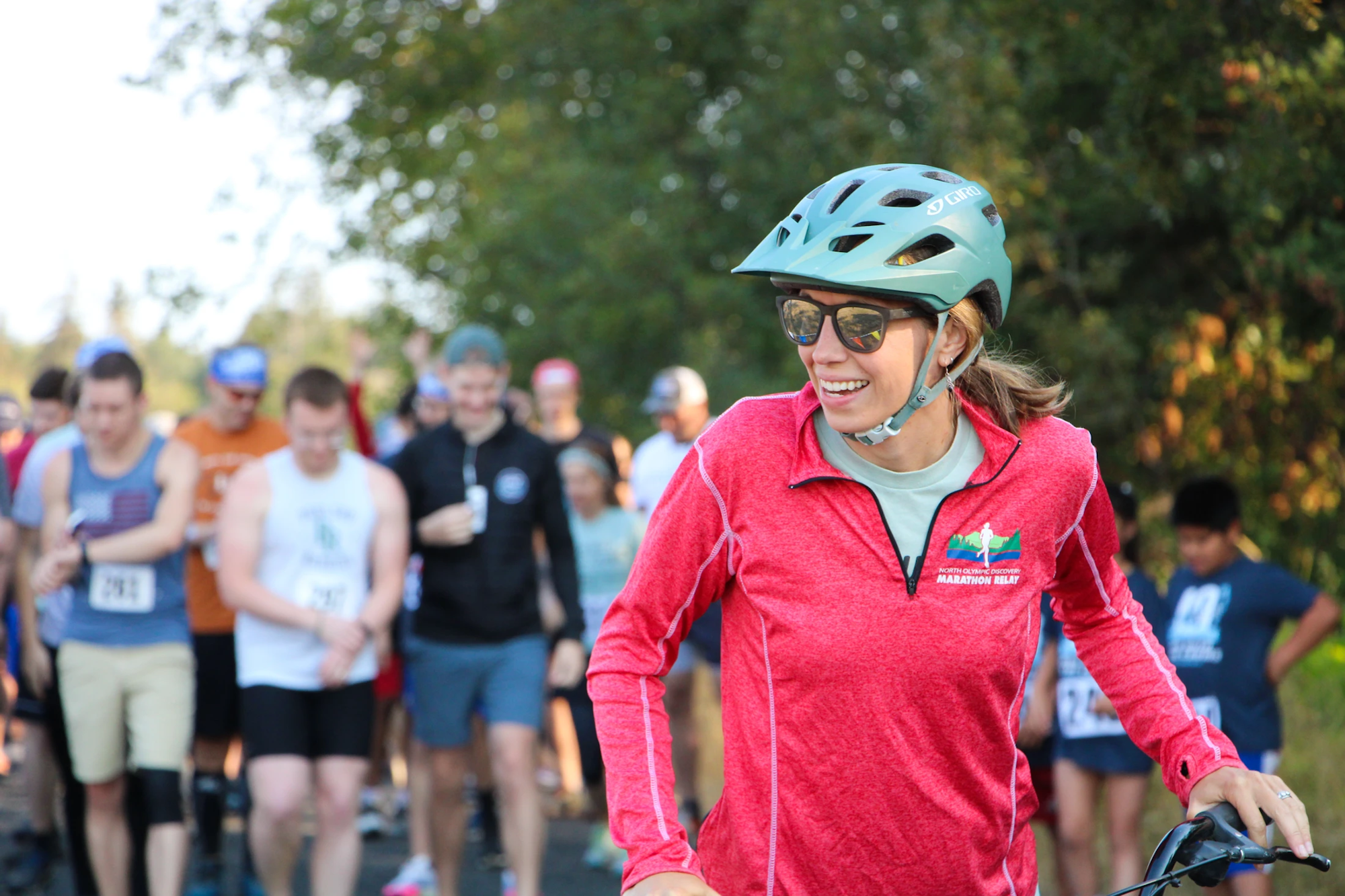 A smiling woman in a red jacket and helmet, holding a bike, with runners in the background.