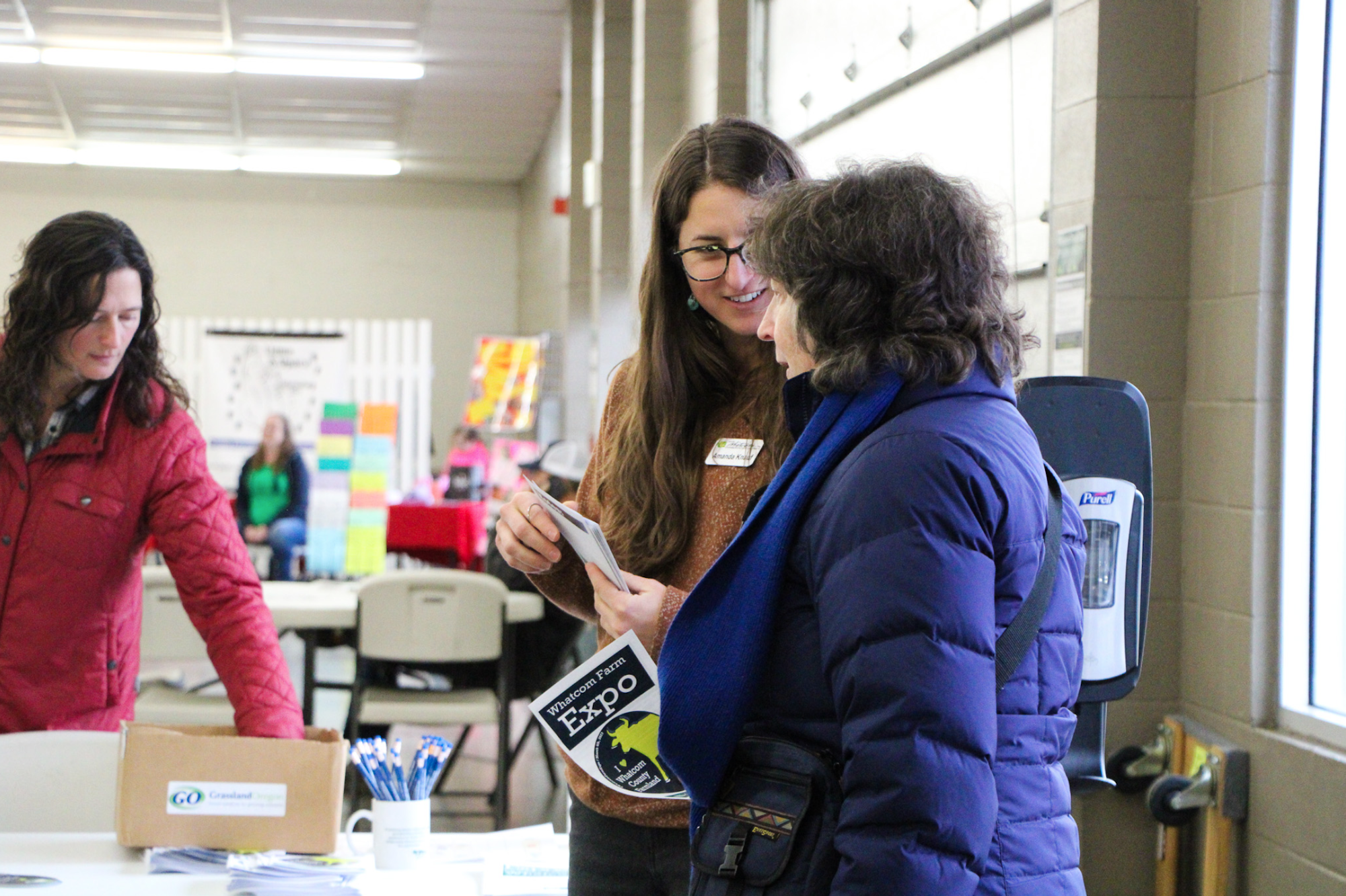 Two people talking at an event table, holding a brochure titled "Whatcom Farm Expo." Another person is looking at papers on the table.