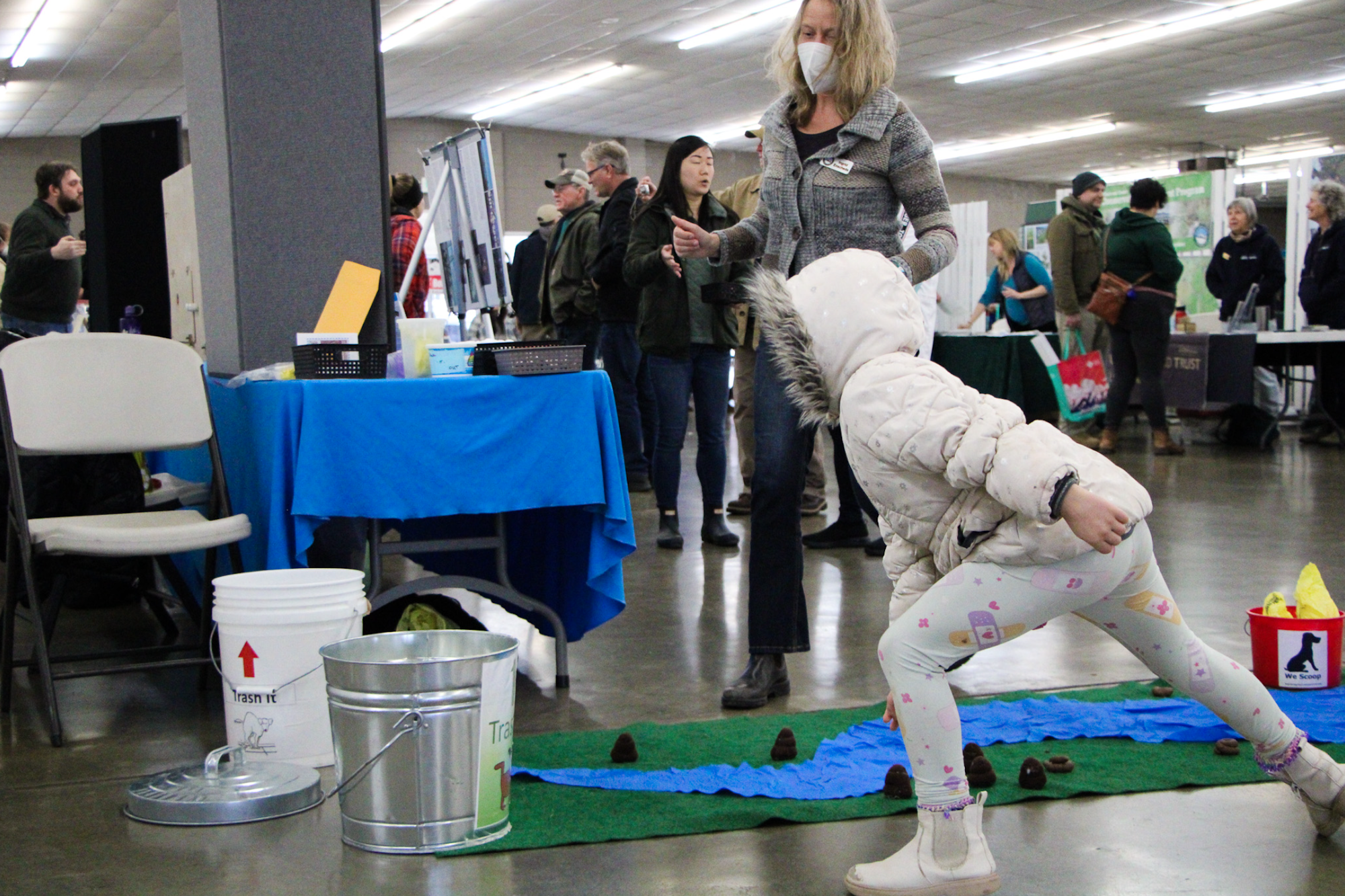 A child plays a game indoors, hopping over items on the floor while people and booths are in the background.