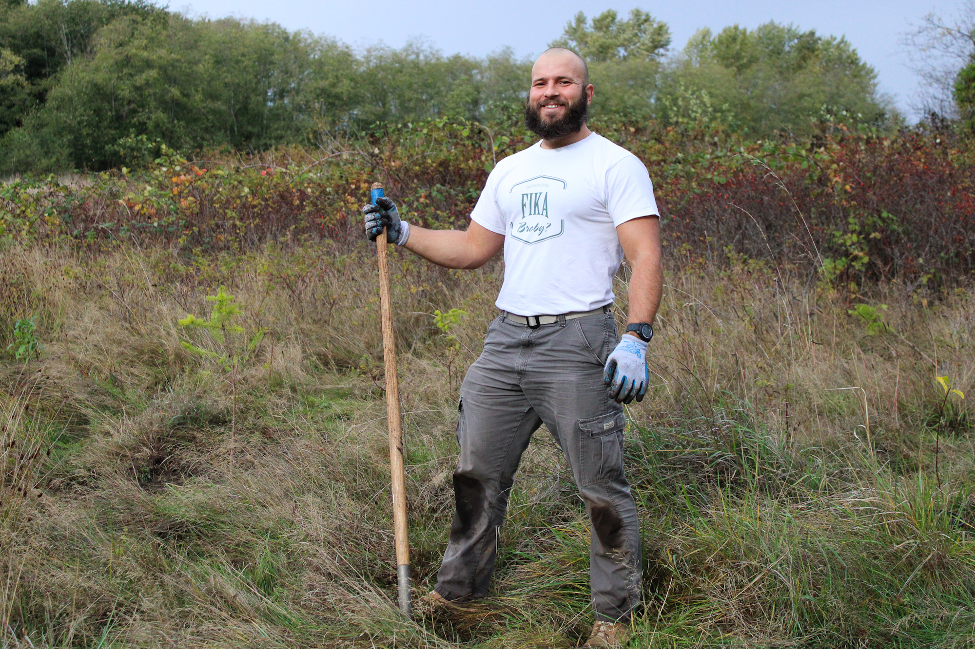 A man standing in a field holding a shovel, wearing a white t-shirt and gloves.