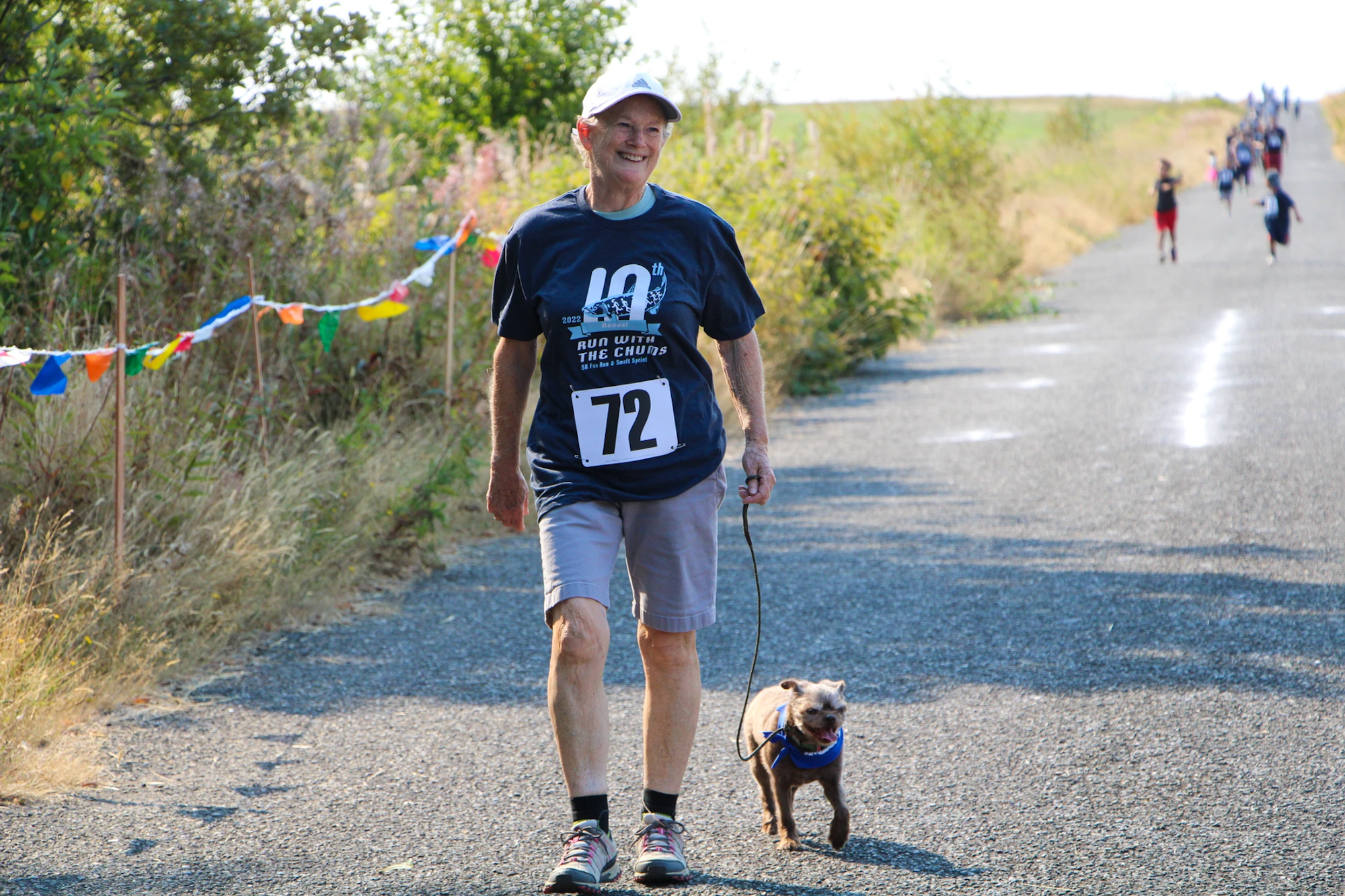 A person in a race bib walks a small dog on a path, with colorful flags in the background.