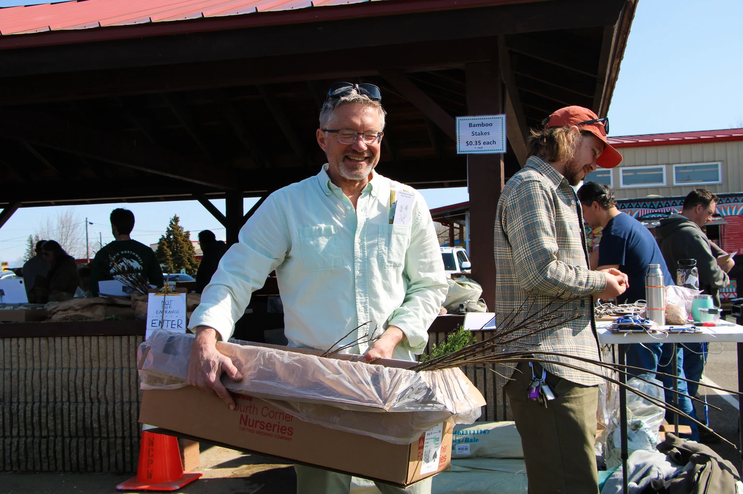 Man holding a cardboard box with tree branches, standing at an outdoor market or fair.