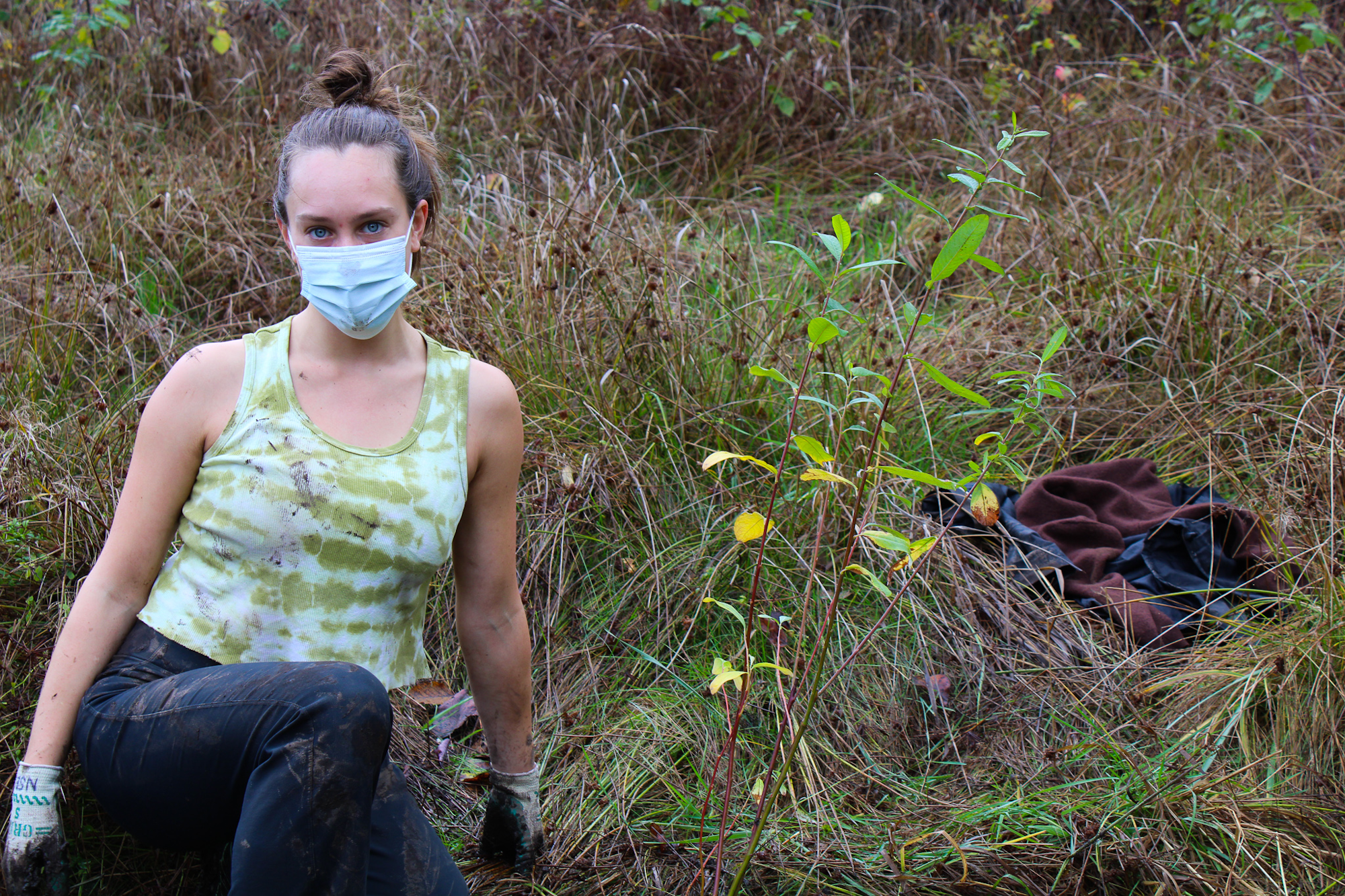 A person wearing a mask and gloves sits in a grassy field with plants and clothing scattered nearby.