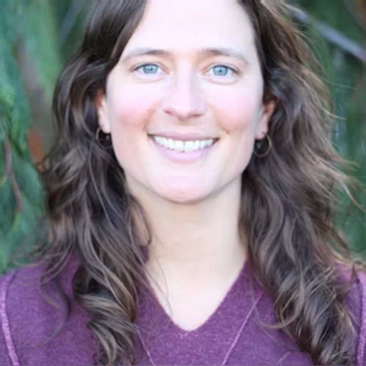 A person with long, wavy hair is smiling, wearing a purple top, with a blurred green background.