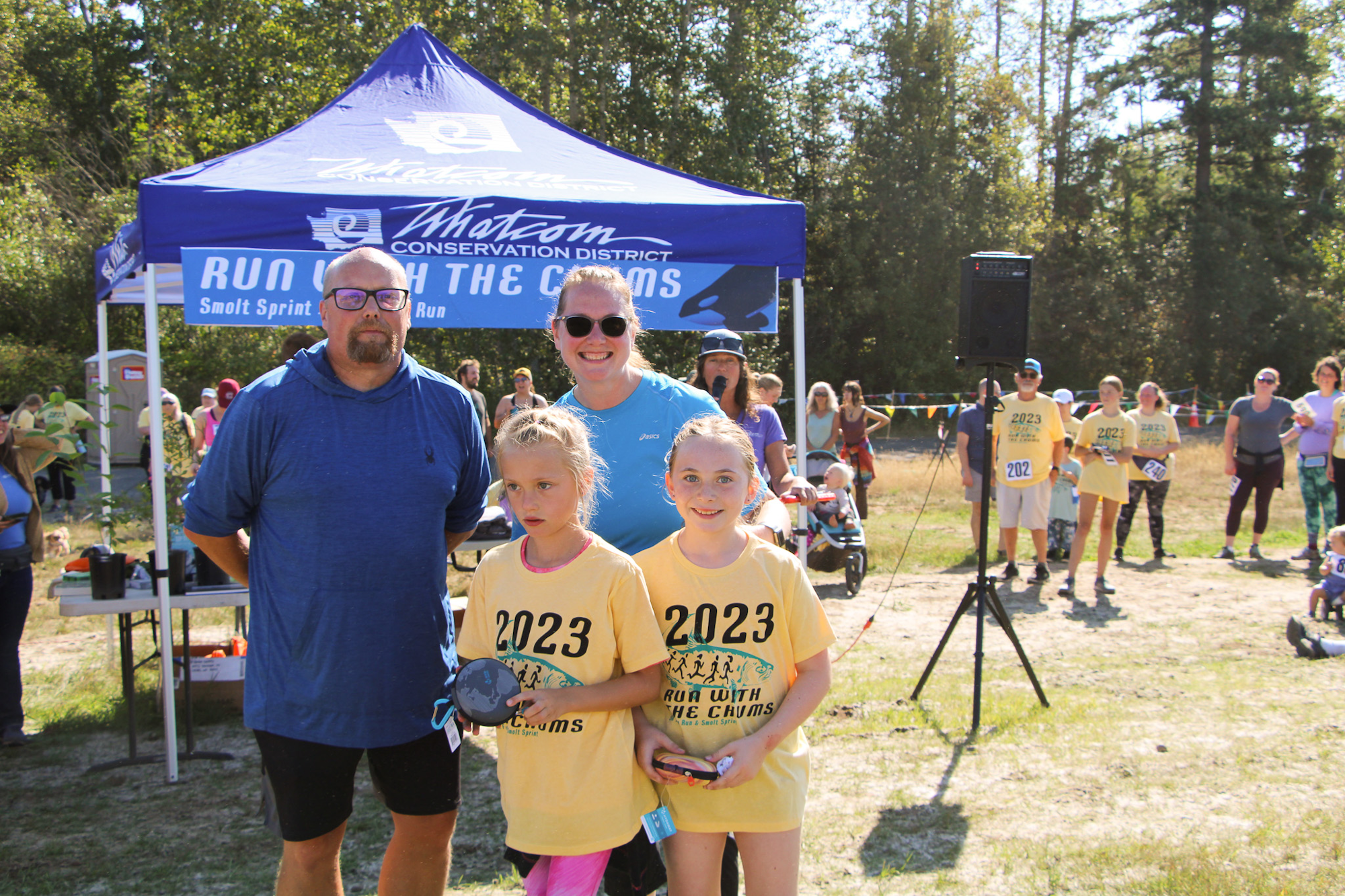 A family poses in front of a "Run with the Chums" tent at an event. The children wear matching 2023 event shirts.