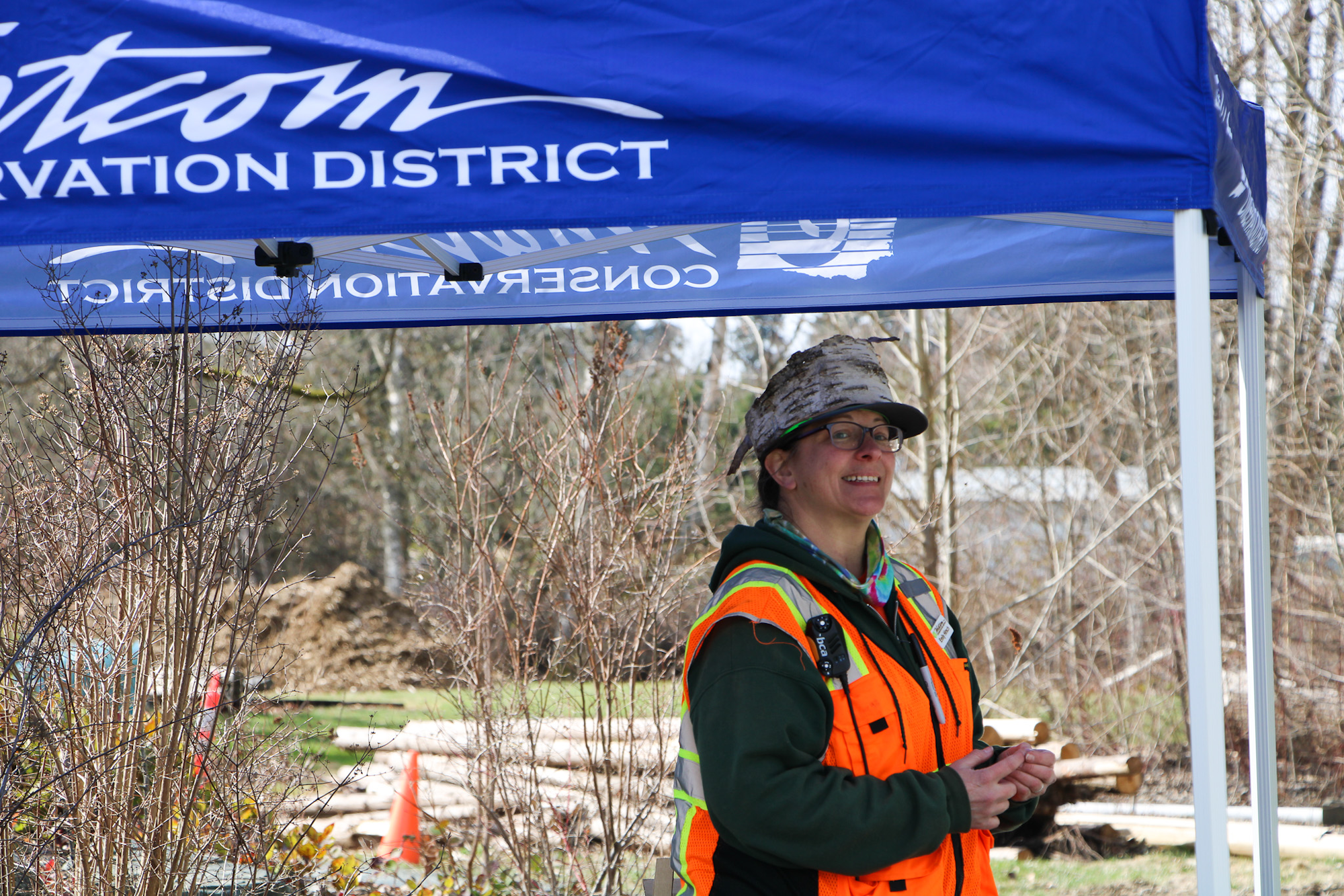 Person in safety gear under a blue tent labeled "CONSERVATION DISTRICT," outdoors with trees and construction materials.