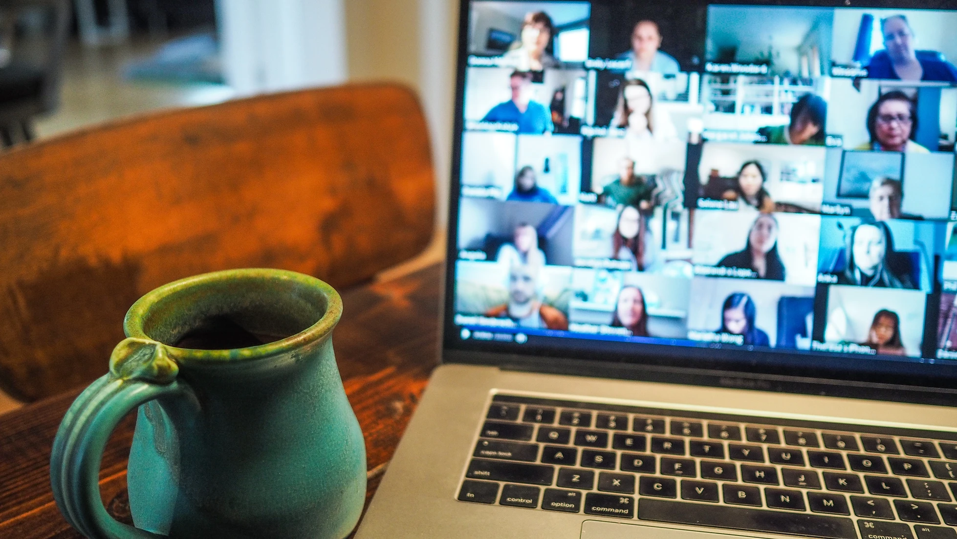 A laptop displaying a video call with many participants and a green mug on a wooden table.