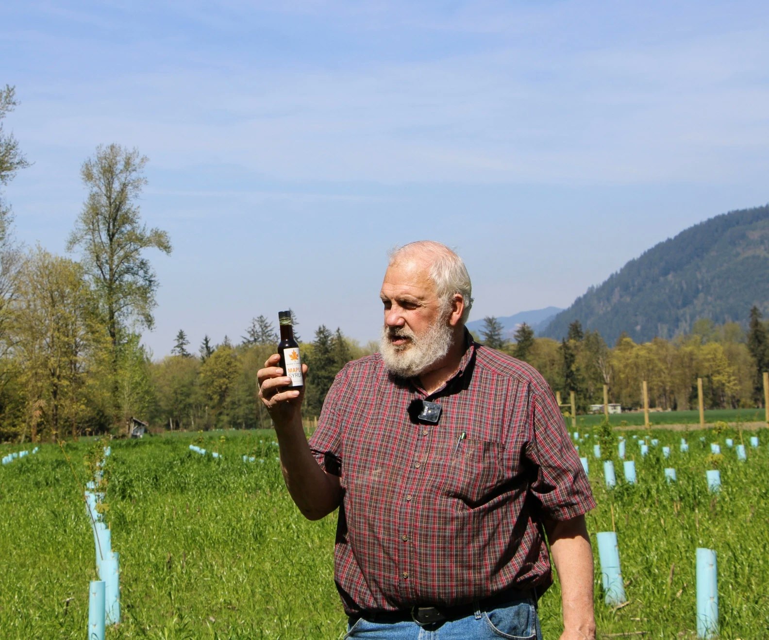 A man in a field holds a small bottle; trees and mountains are in the background.