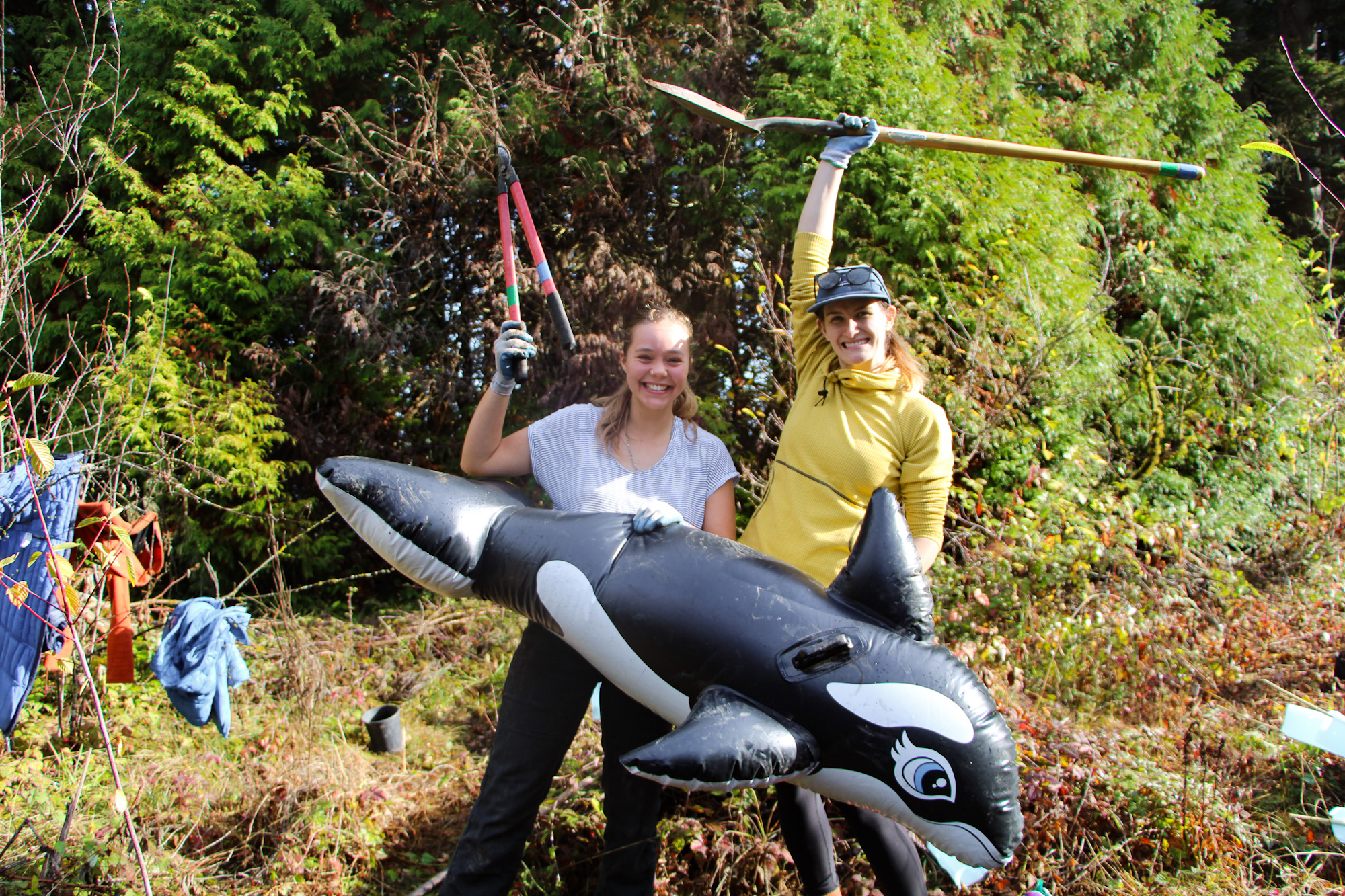 Two people outdoors, smiling, holding an inflatable orca and gardening tools, surrounded by greenery.