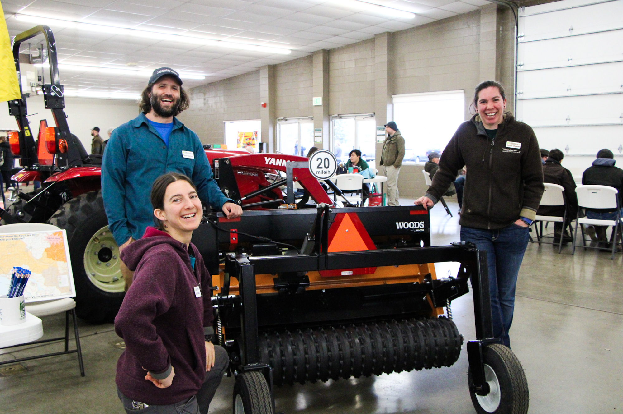 Three people smiling near farm equipment at an indoor event.