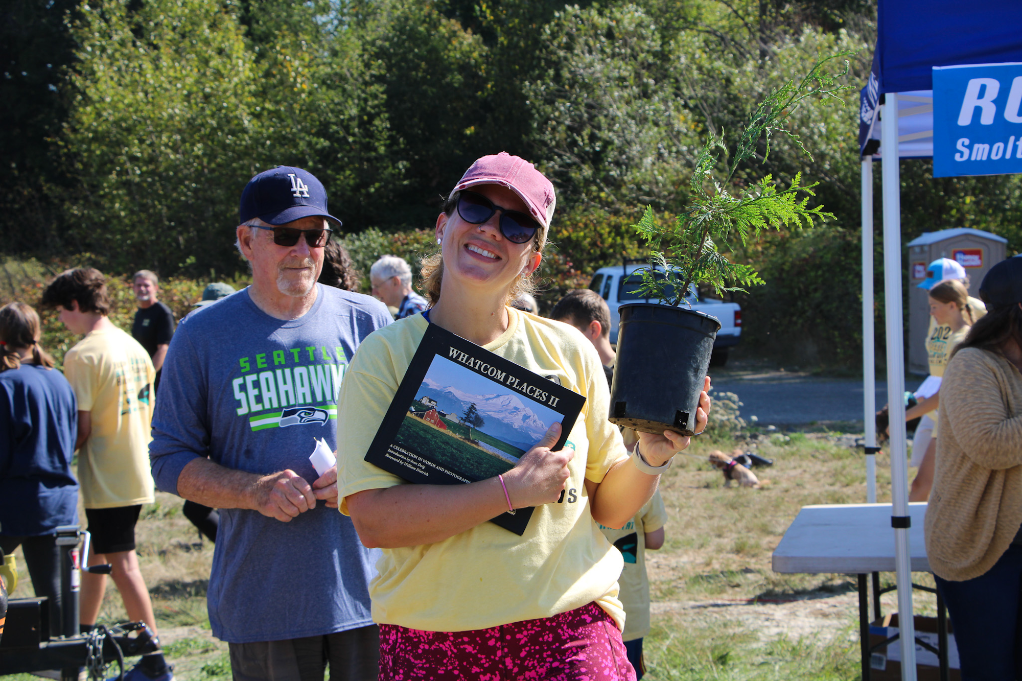 A woman smiles holding a book and a potted plant at an outdoor event. People are gathered in the background.