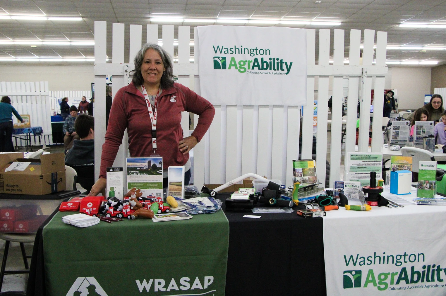 A woman stands at a Washington AgrAbility booth displaying farming tools and brochures at an indoor event.