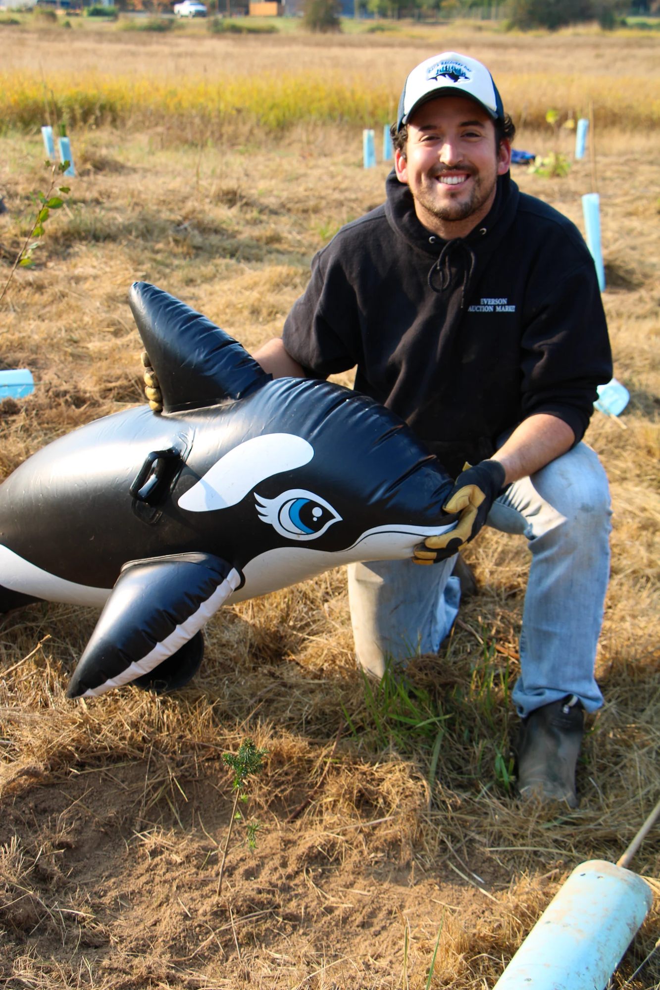 A person kneels in a field, smiling, holding an inflatable orca.