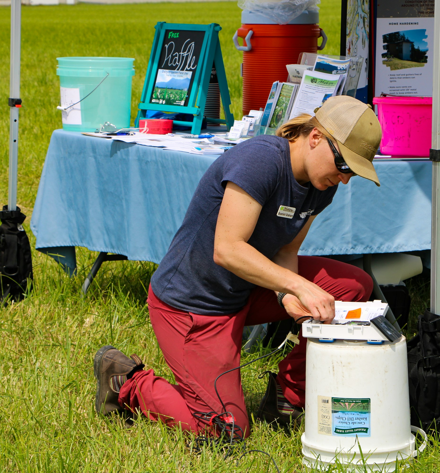 A person is kneeling by a booth with a raffle sign, working with documents and a device on a white bucket in a grassy area.