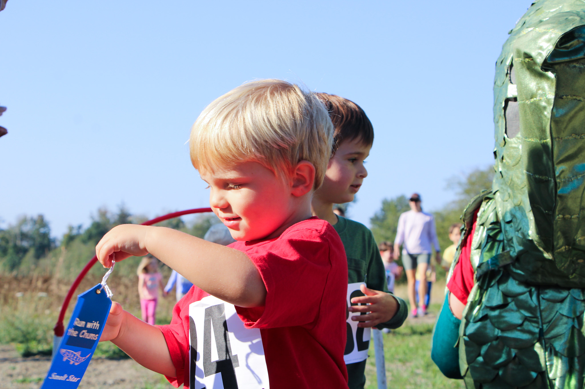 Two young children with number tags, one holding a blue ribbon, outdoors with others in the background.