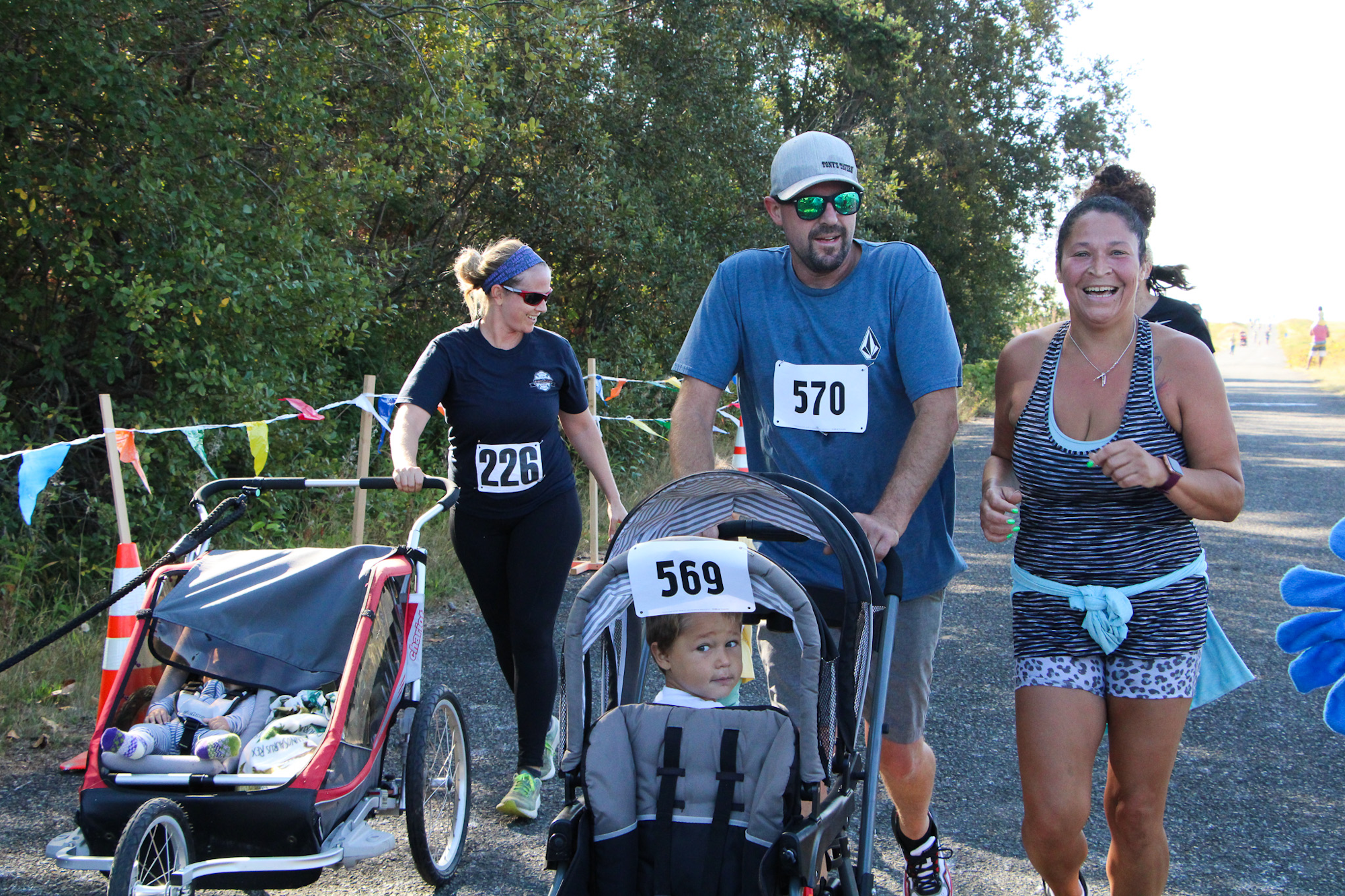 People jogging in a race, one pushing a stroller, another with a buggy, wearing numbered bibs, smiling outdoors.