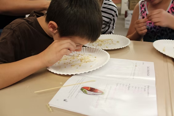 A child examining a paper plate with a magnifying glass, near an open book on a table, with other children nearby.