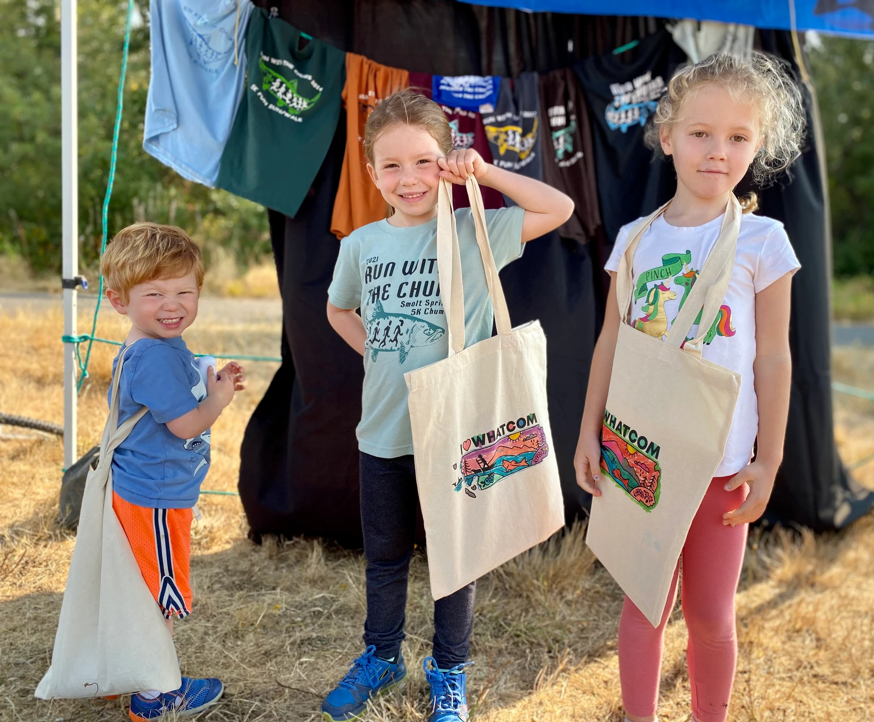 Three children smiling and holding tote bags with colorful designs in front of a display of T-shirts.