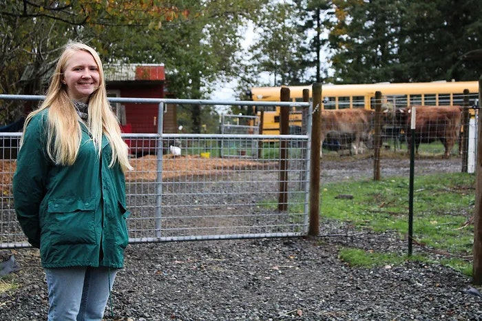 A person stands smiling in front of a gate with a farm scene and a yellow school bus in the background.