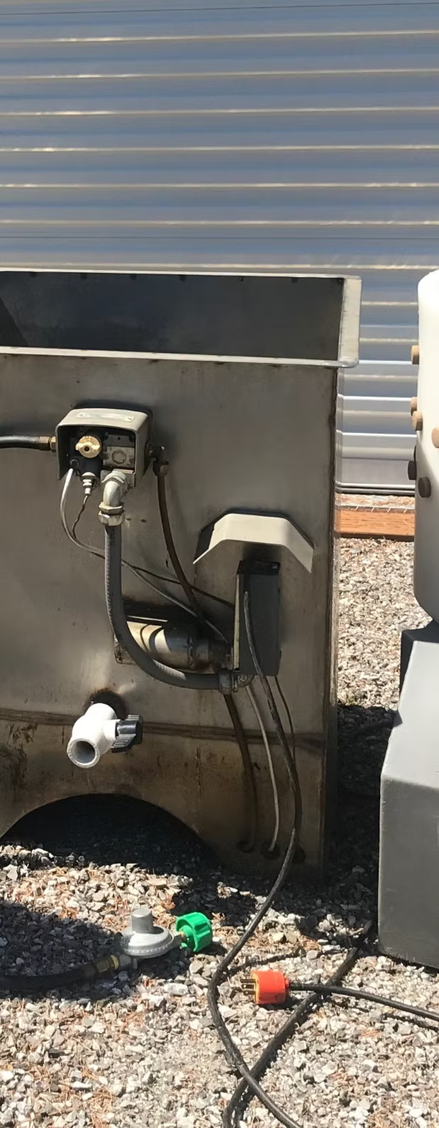 Outdoor equipment setup with metal tank, hoses, valves, and cables on gravel ground near corrugated metal wall.