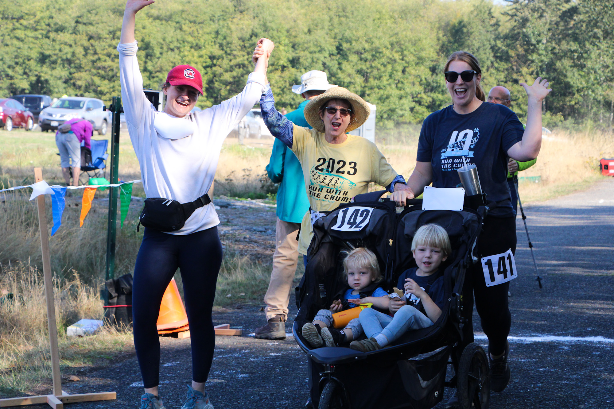 People celebrating at a race finish line; two women, a stroller with kids, race numbers, and colorful flags.