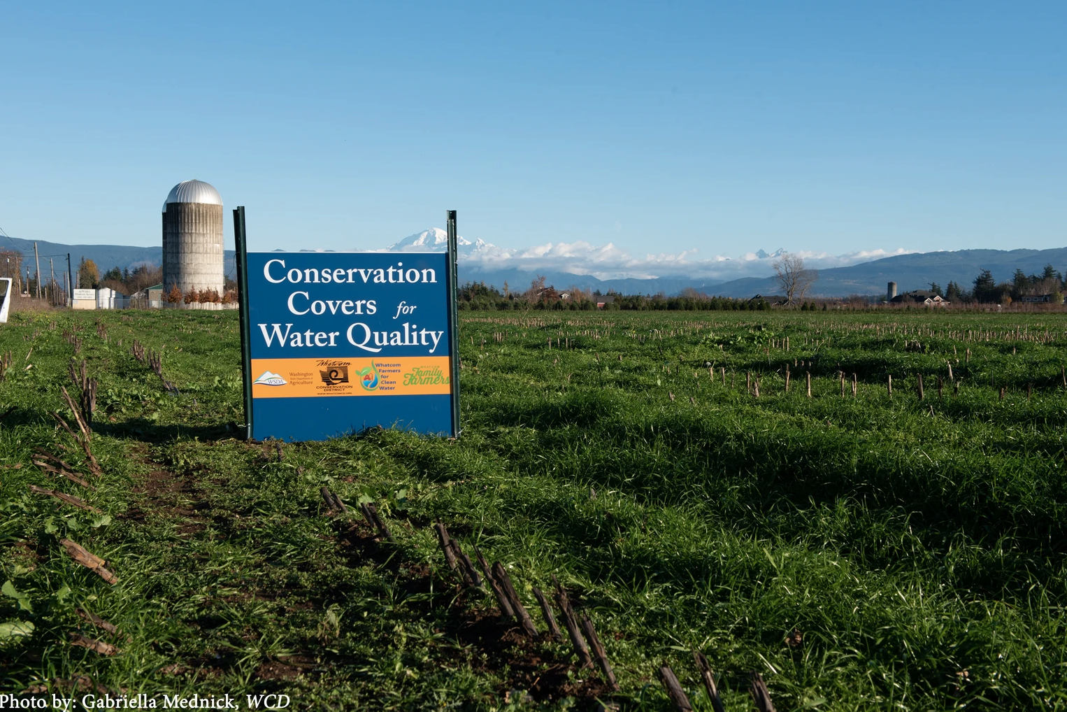 A field with a sign that reads "Conservation Covers for Water Quality" against a backdrop of mountains and a silo.
