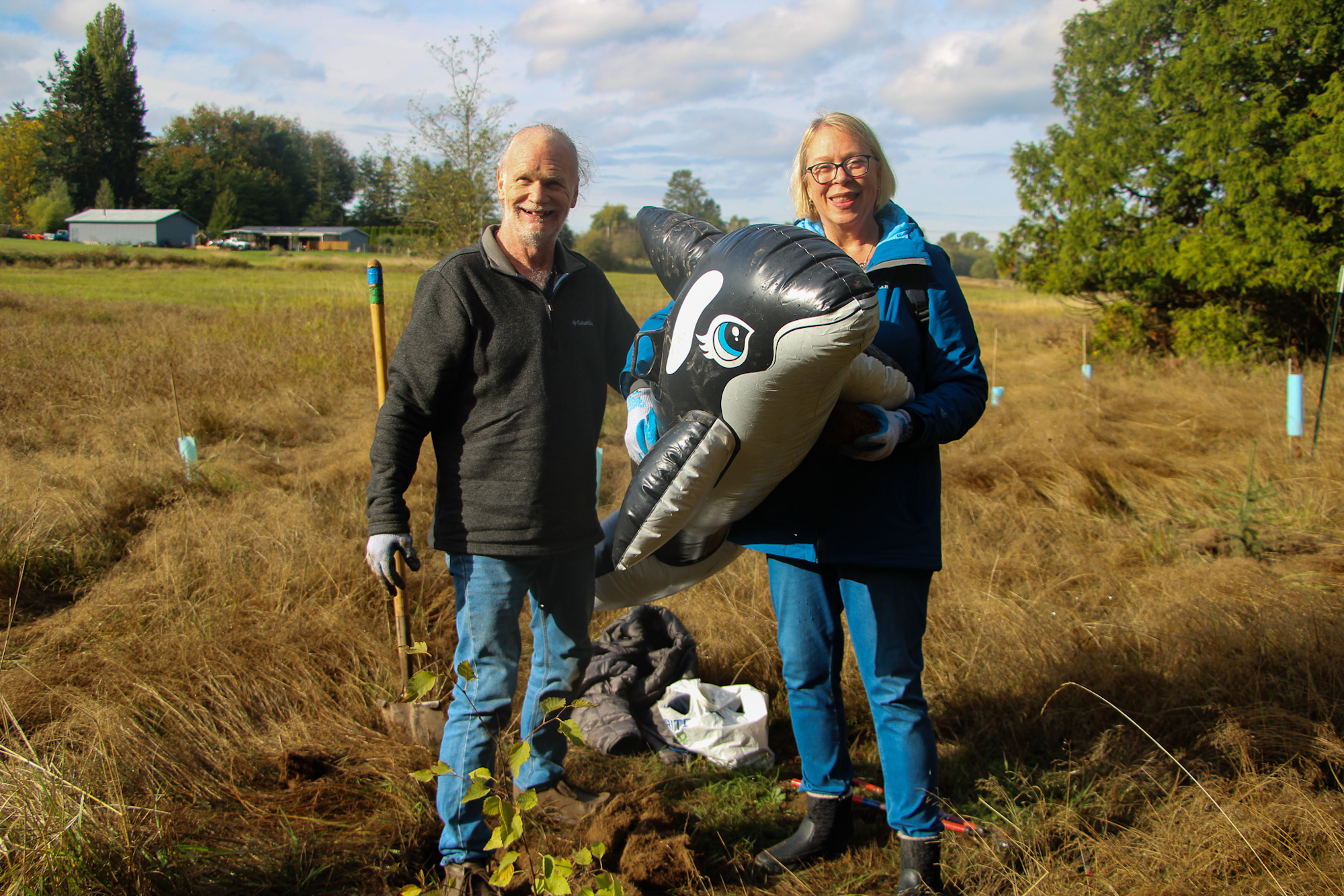 Two people standing outdoors, one holding a shovel and the other an inflatable orca.