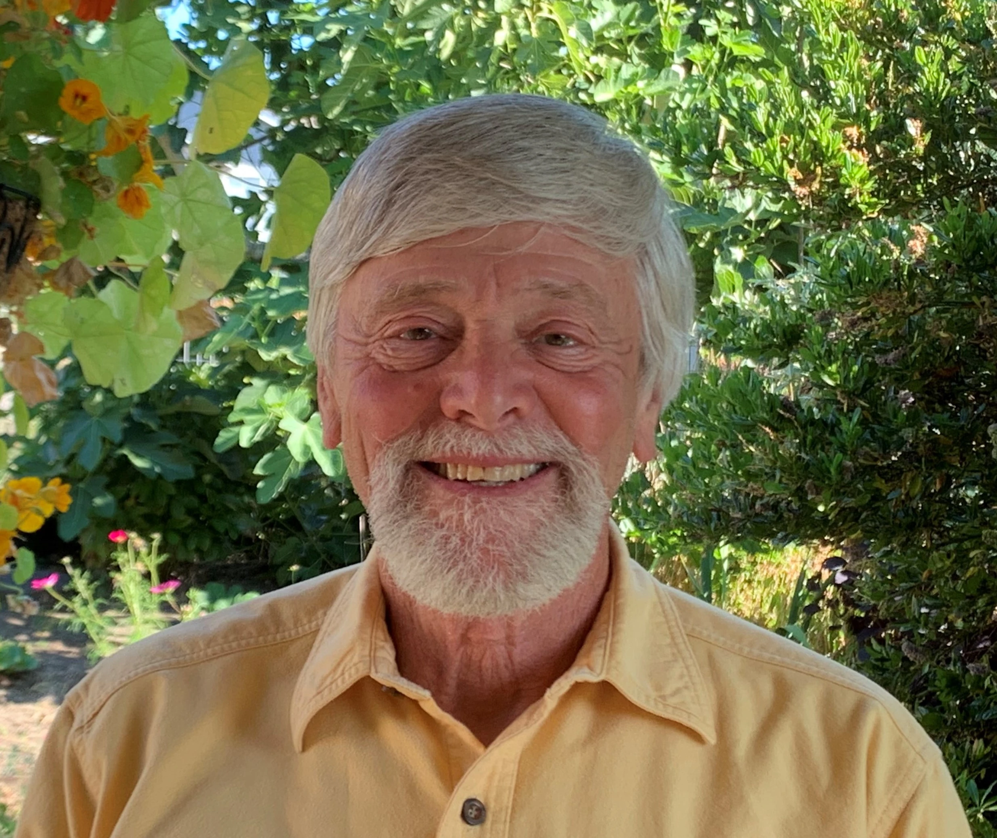 A smiling man in a yellow shirt stands in front of lush green foliage with orange flowers.